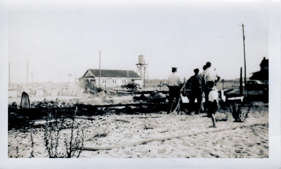 Black and white photograph of men with a hose among burned debris, with the dance hall and the water tower behind them