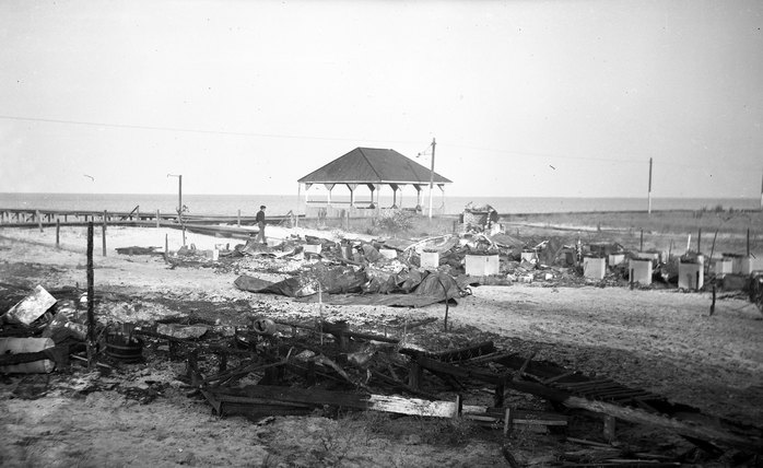 Black and white photograph of burned cottage debris on the sand with the pavilion and the Delaware Bay in the background